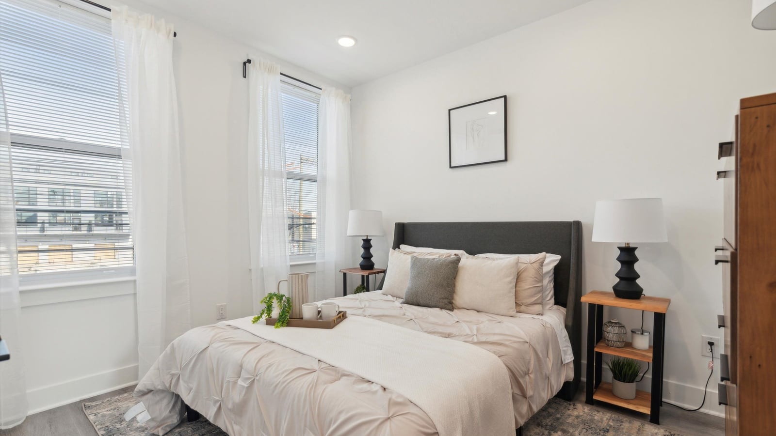 Staged bedroom with a bed, two bedside tables with lamps, two windows with blinds and sheer curtains, white walls and gray plank flooring
