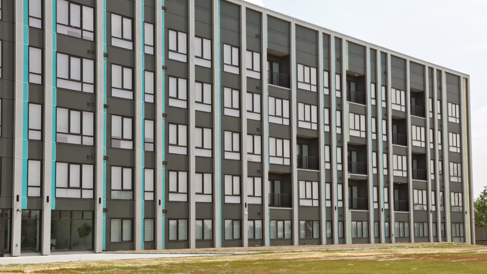 Six-story apartment building with dark gray panels, pale vertical piers, turquoise accent strips, rows of windows and recessed balconies, seen across a patch of grass