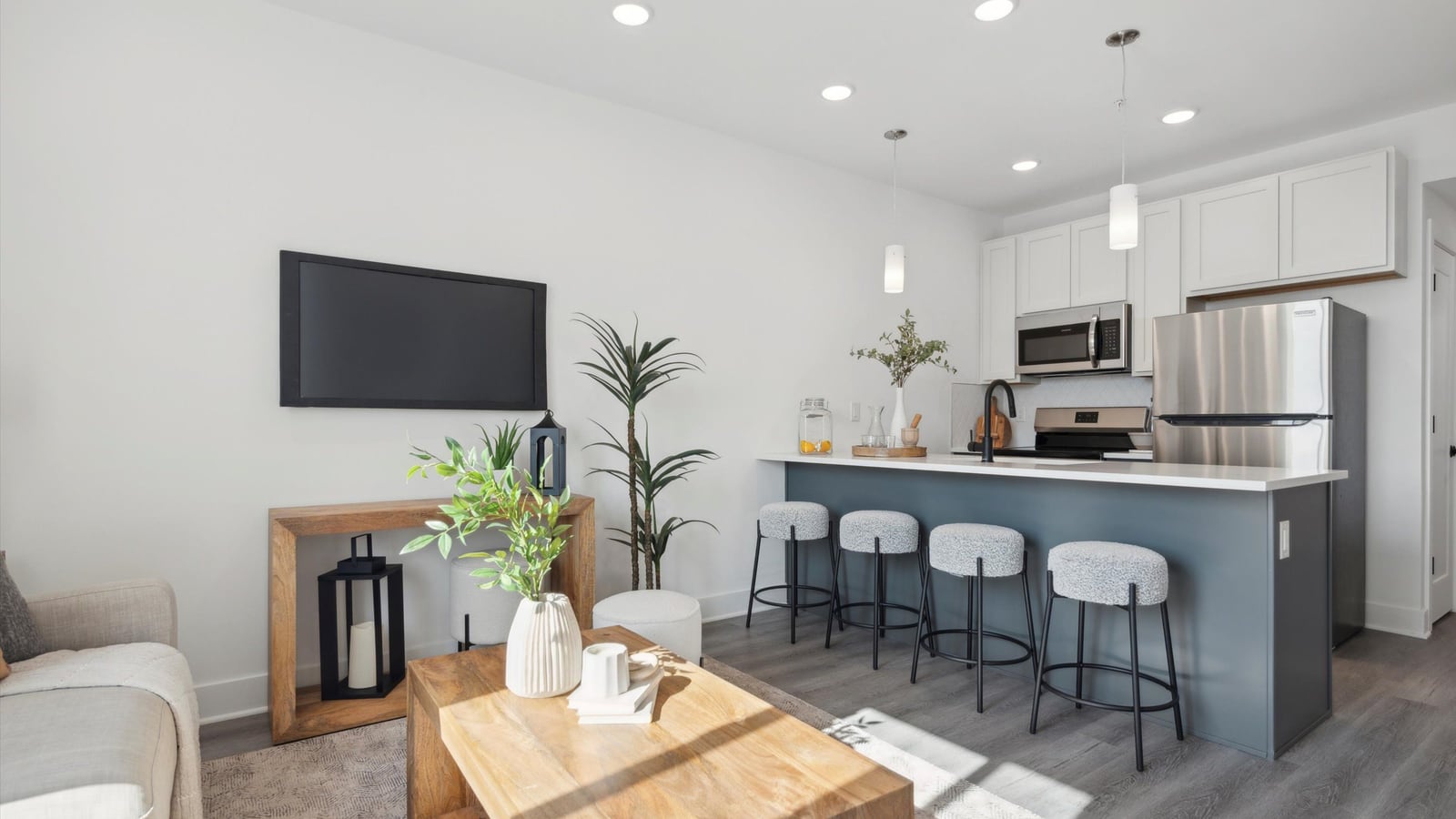 Furnished living area and kitchen with white upper cabinets, a blue-gray island with four stools, a stainless steel refrigerator, range and microwave, a television on the wall and gray plank flooring