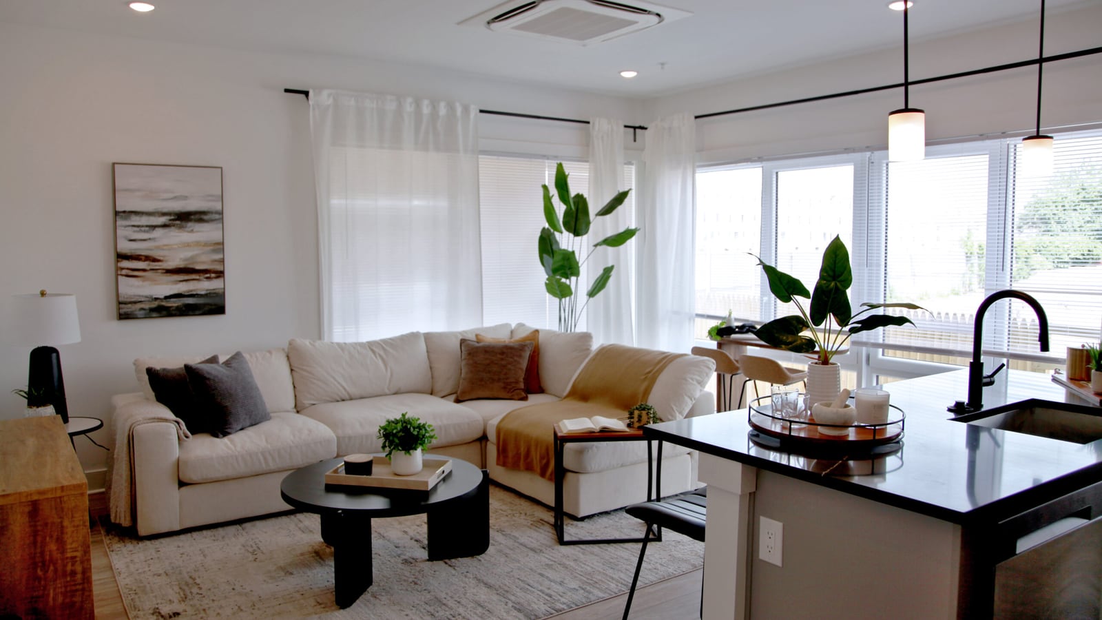 Furnished living room with a pale sectional sofa, an oval black coffee table, a kitchen island with a black faucet, tall windows with white curtains and an air conditioning unit set in the ceiling
