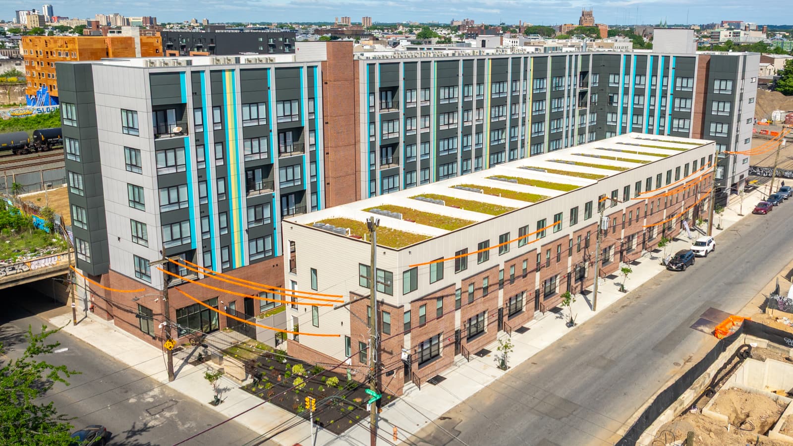 Aerial photograph of a six-story apartment building with dark gray and pale panels and blue and yellow vertical stripes, behind a row of three-story brick and white buildings with planted roofs along a street, with rail tank cars on tracks at the left