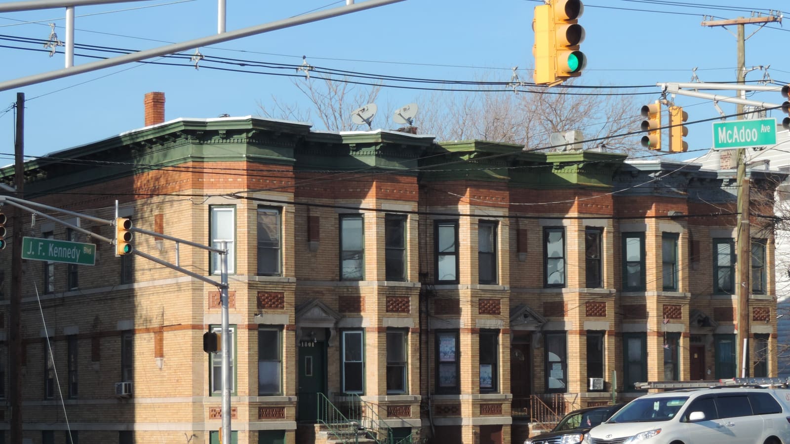 A row of two-story yellow and red brick row houses with green cornices at a street corner, with street signs for J. F. Kennedy Blvd and McAdoo Ave