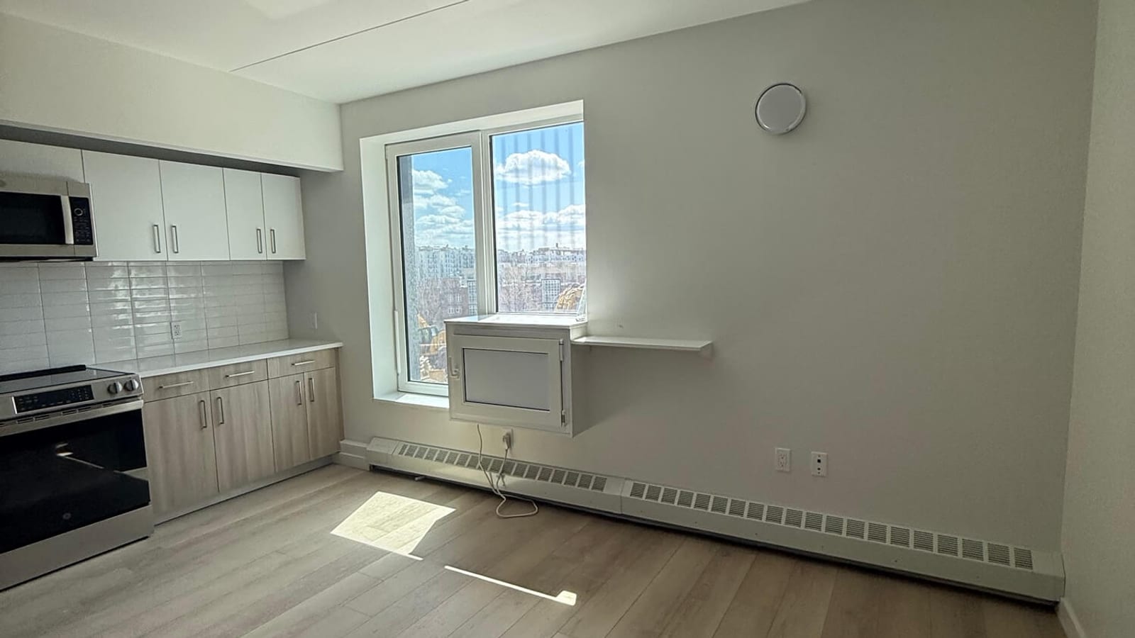Photograph of an empty room with pale walls and wood-look plank flooring, a two-pane window looking out on apartment buildings and sky, a white box-shaped cover on the wall below the window, a baseboard unit along the wall, and at the left a kitchen run with white upper cabinets, a tile backsplash, a microwave and an electric range
