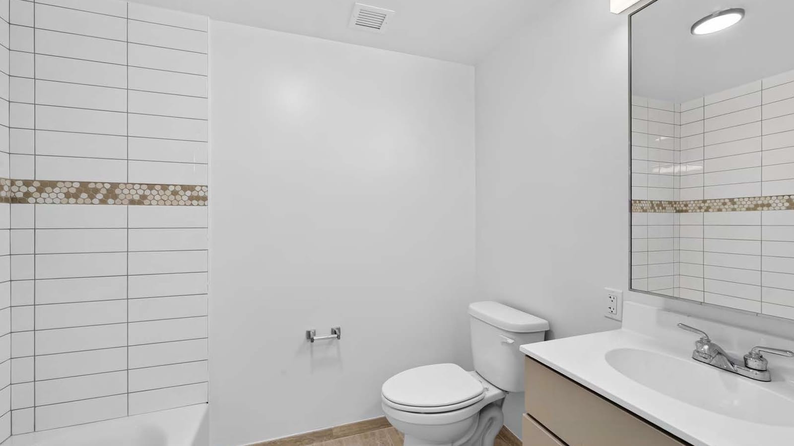 Bathroom with a white bathtub and white stacked wall tile crossed by a band of tan and white hexagonal mosaic, a white toilet, a vanity with a white top and a beige cabinet, a mirror with a light bar above it and tan floor tile