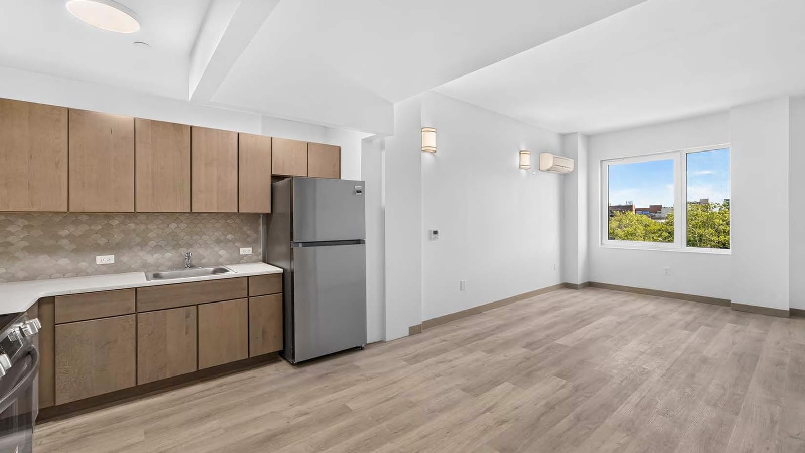 Empty apartment interior with a kitchen along the left wall, wood-tone upper and lower cabinets, a white counter with a steel sink, a scalloped tile backsplash, a stainless steel refrigerator and the edge of a range, opening onto a room with pale wood-look flooring, white walls, wall sconces, a wall-mounted heating and cooling unit and a large window looking out on trees