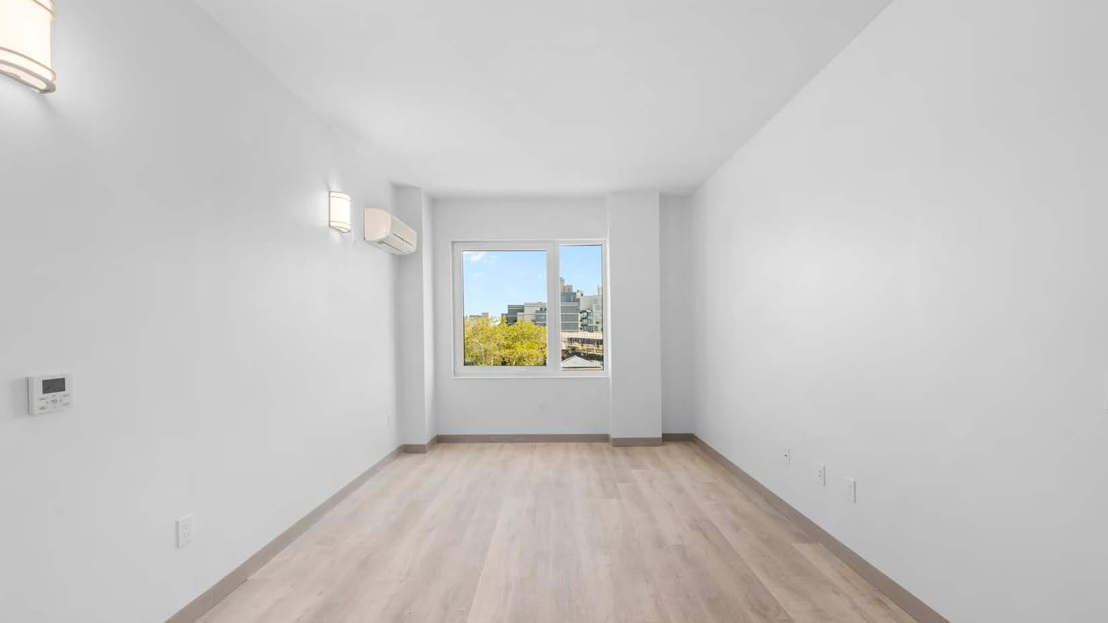 Empty rectangular room with white walls and pale wood-look flooring, two wall sconces, a wall-mounted heating and cooling unit beside a two-pane window that looks out on trees and mid-rise buildings, and a small wall controller at the left