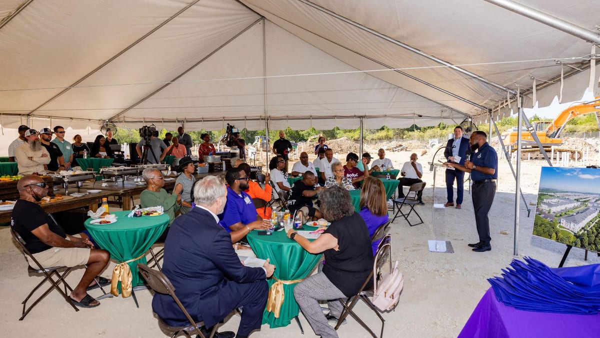 People seated at tables under a white tent listening to a speaker beside an easel holding the building rendering, with an excavator on bare ground behind them