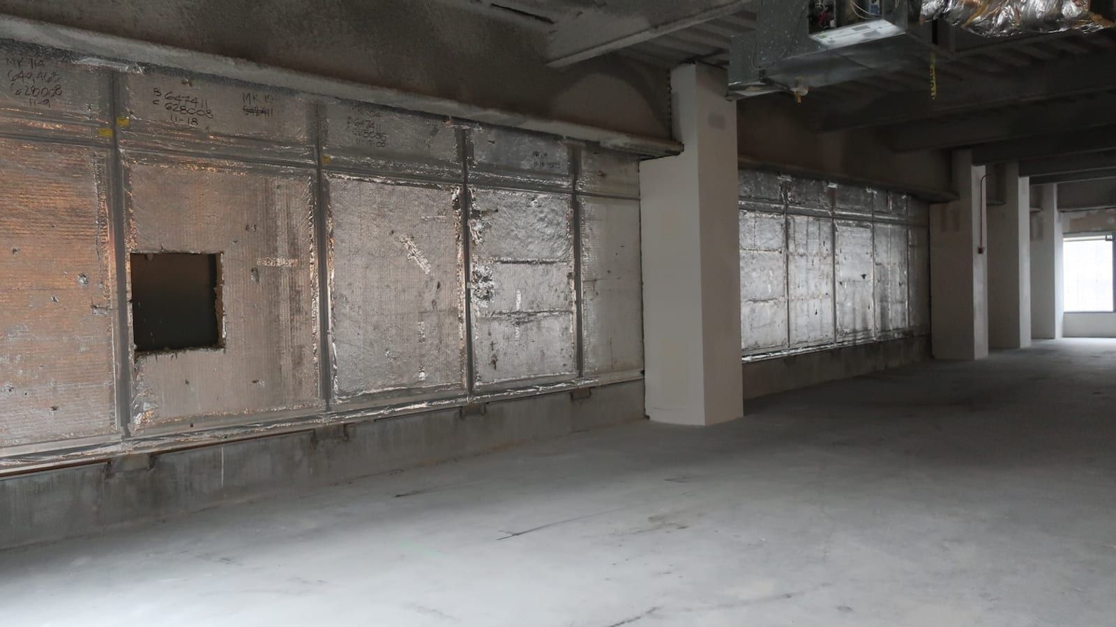 Photograph of an empty office floor with bare concrete floor, foil-wrapped insulation panels along the outer wall, one panel cut open, white columns, exposed ductwork overhead and daylight from windows at the far end