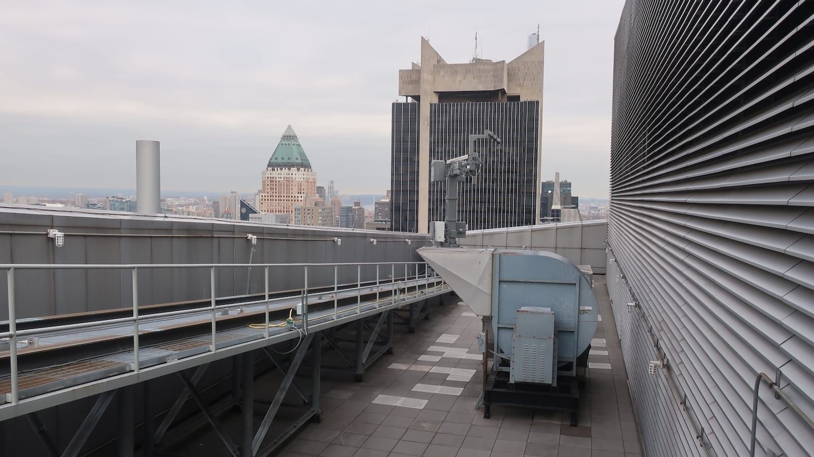 Photograph of a setback roof with a raised metal walkway, a large grey air handling unit and a wall of grey louvers, with Midtown towers and a green pyramid roof in the distance