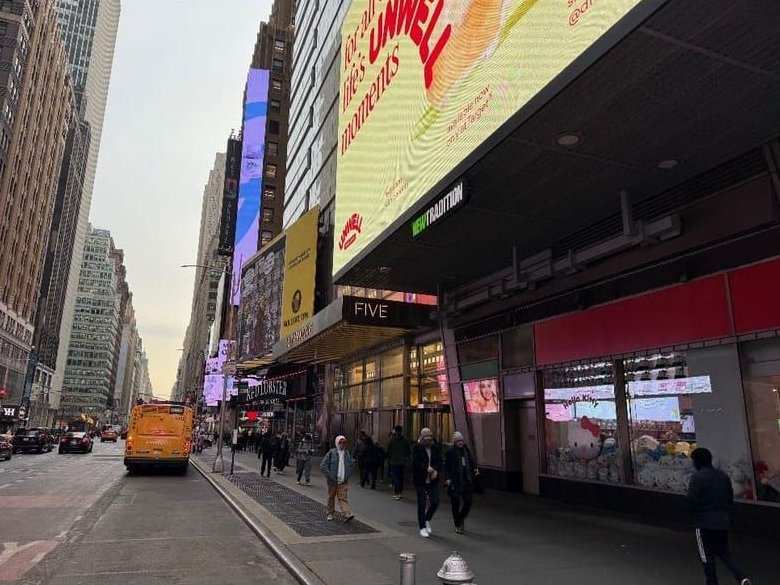 A busy Seventh Avenue sidewalk along the base of the tower, with a large yellow LED advertising screen over the entrance, a sign reading FIVE, shop windows and a yellow bus in the street