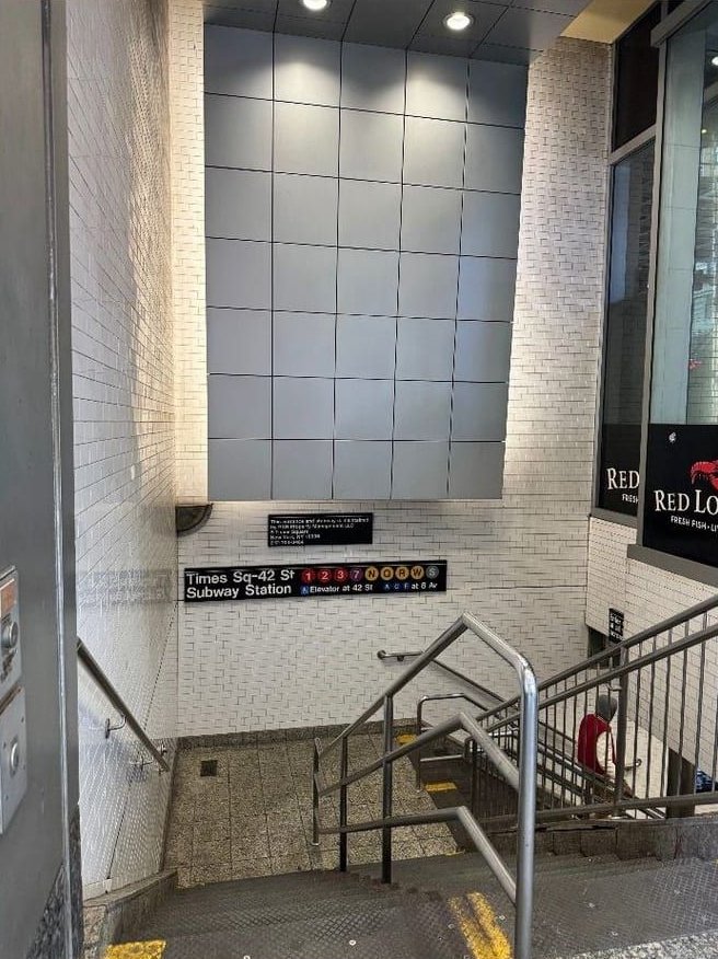 A stairway down to the subway inside the building's base, with a sign reading Times Sq-42 St Subway Station and the line symbols 1, 2, 3, 7, N, Q, R, W and S