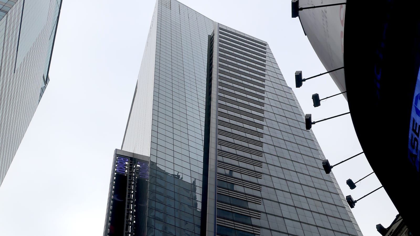 Photograph looking up at a tall glass office tower with a vertical band of grey louvers, a lit purple sign panel near its base on the left, a curved glass building at the right edge with floodlights on arms, and a grey sky