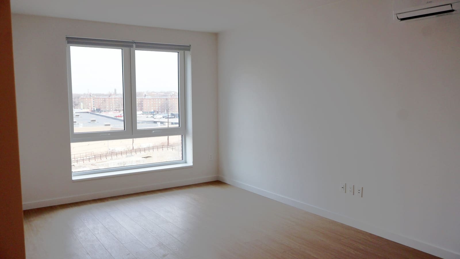 Photograph of an empty room with light wood-look flooring, white walls, a wall-mounted air-conditioning unit and a large two-pane window looking over low rooftops toward brick apartment buildings
