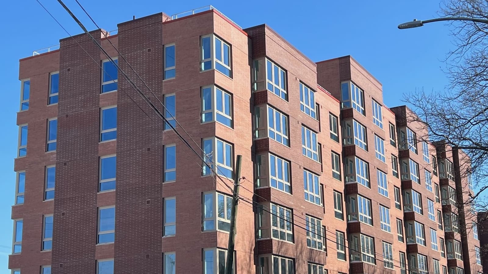 Photograph looking up at a seven-story red brick apartment building with projecting bay windows under a clear blue sky, with utility wires and a wooden pole in front