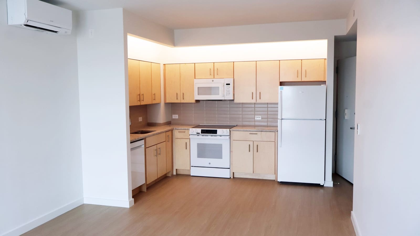 Photograph of an open kitchen with light wood cabinets, a white electric range with a microwave above it, a white dishwasher and refrigerator, grey tile backsplash and a wall-mounted air-conditioning unit