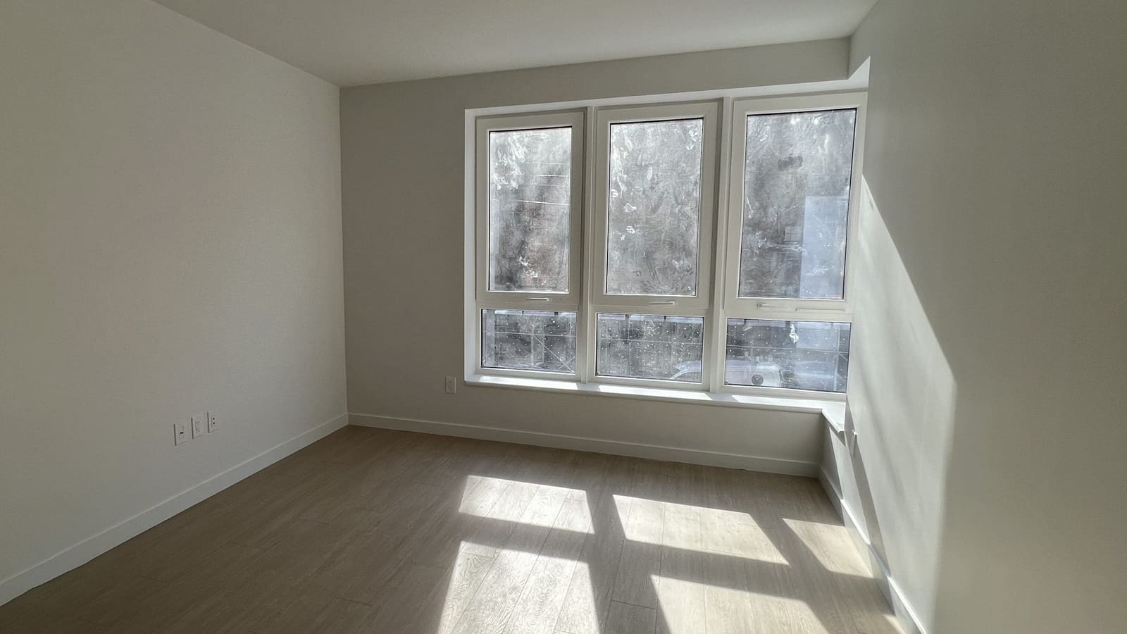 Photograph of an empty room with grey-brown plank flooring, white walls and three tall windows with lower hopper panes, sunlight falling across the floor