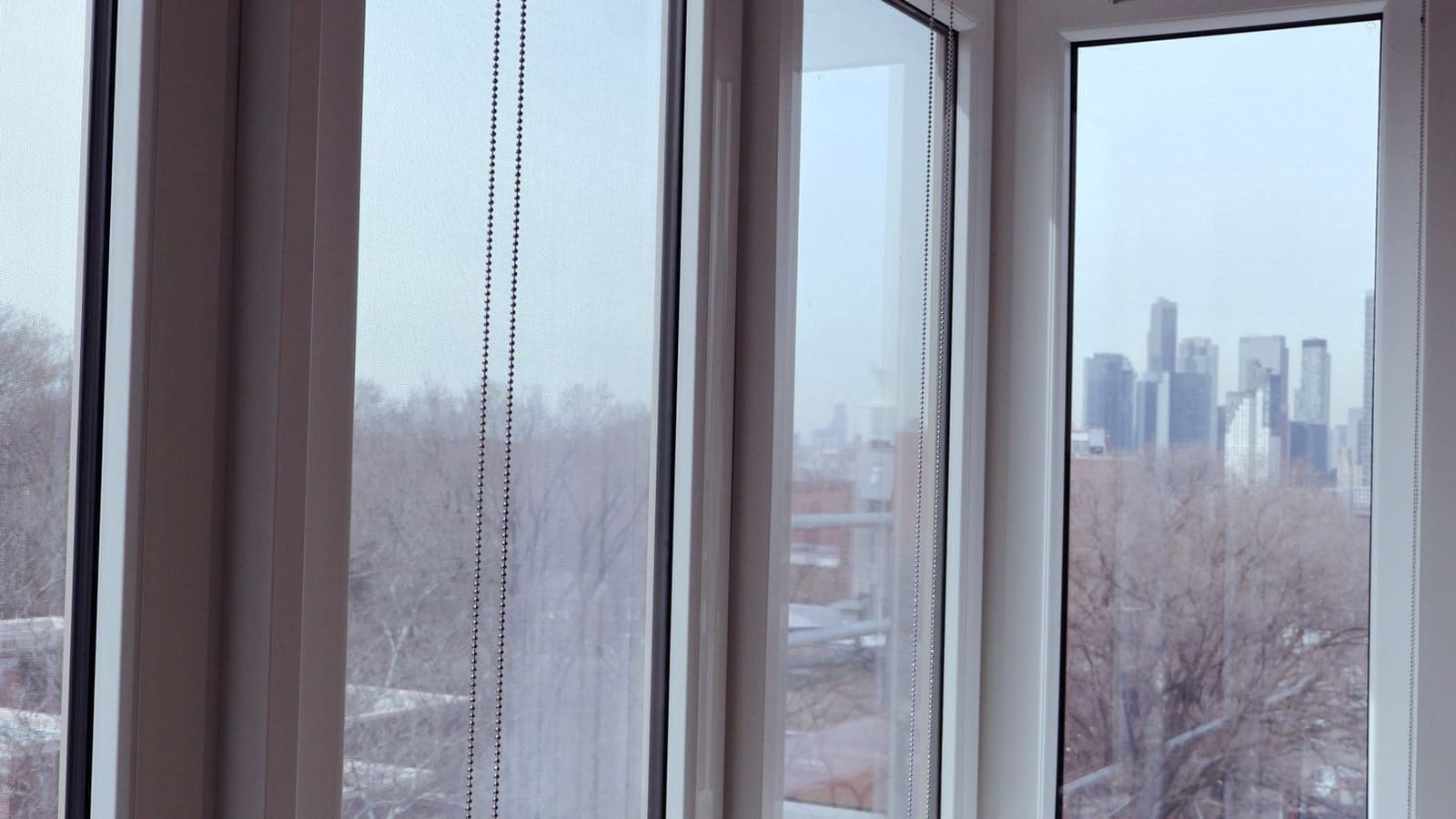Photograph through a corner of floor-to-ceiling windows with roller-shade chains, showing bare treetops and a cluster of distant high-rise towers under a hazy sky