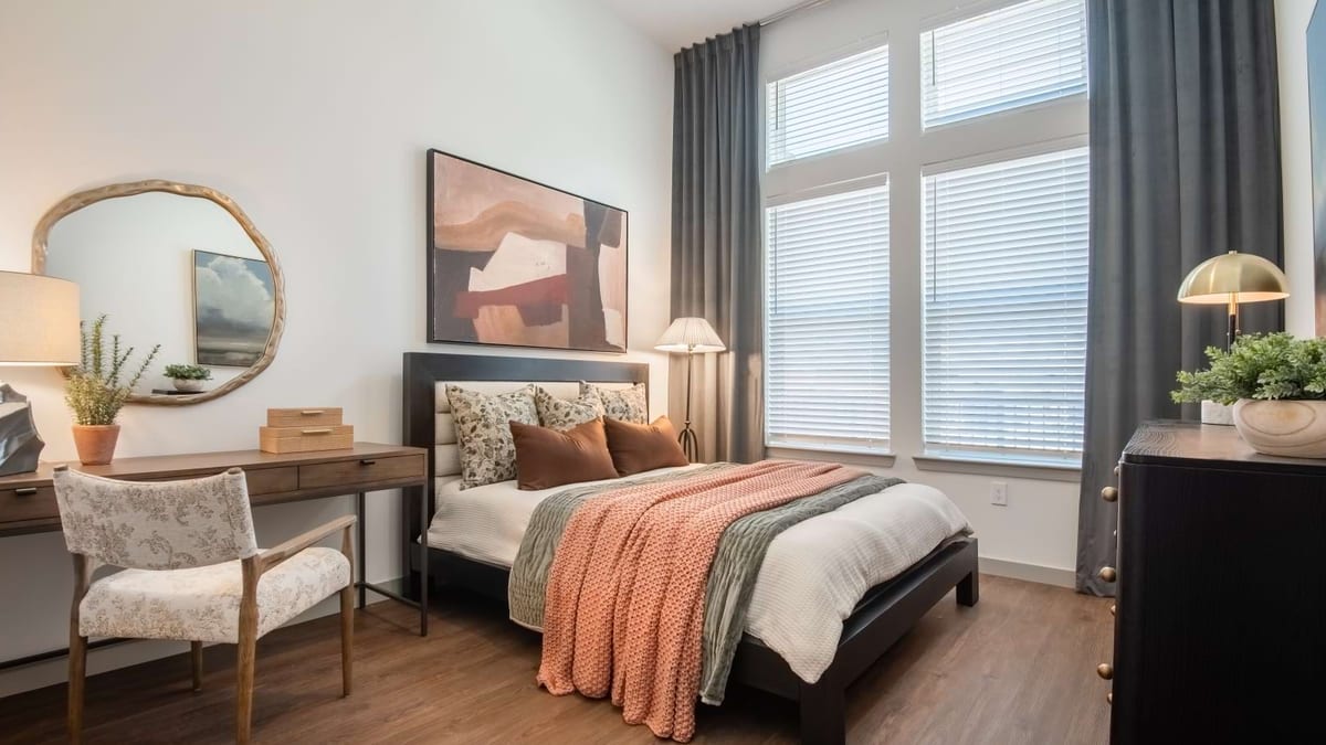 Bedroom with a made bed, an orange blanket, a desk by tall windows and a round mirror
