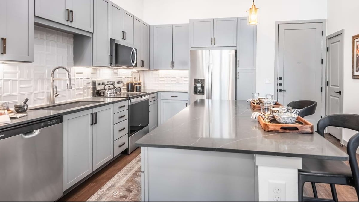 Kitchen with grey cabinets, a white tile backsplash, stainless-steel appliances and a long island with a dark counter