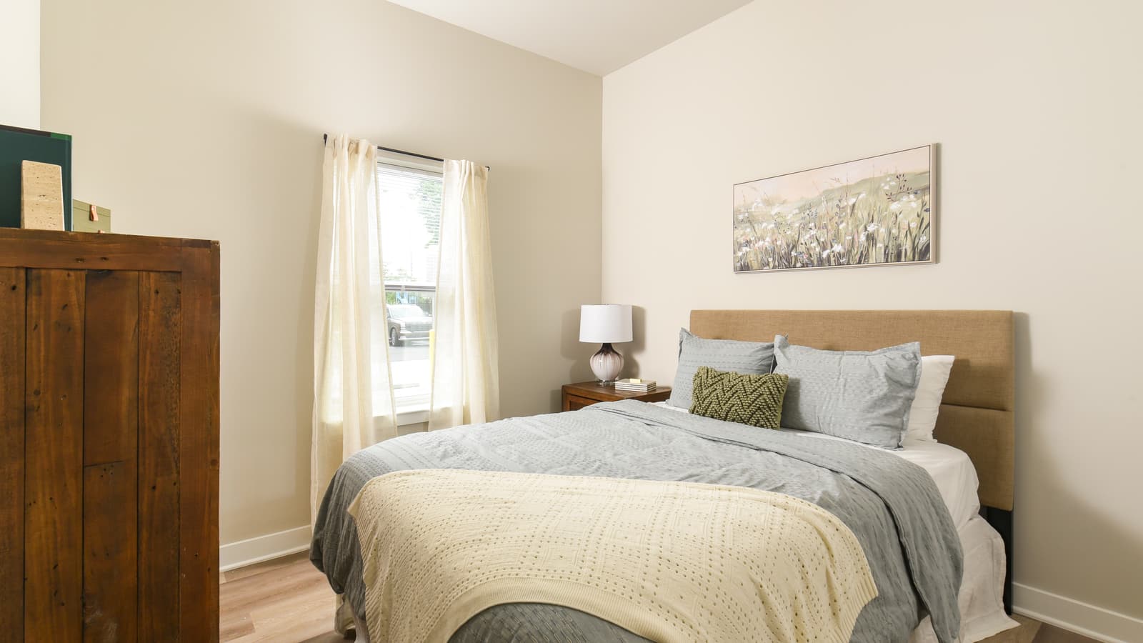A bedroom with a made bed, a wooden dresser, a framed print and a window with white curtains