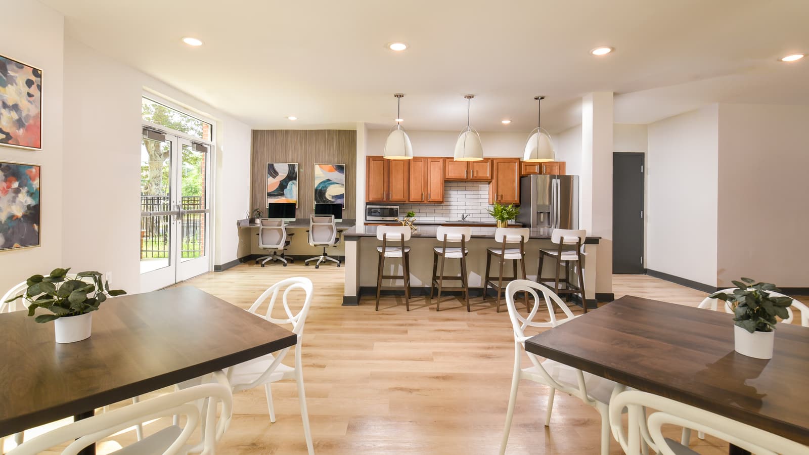 A community room with dining tables and white chairs, a kitchen island with bar stools under pendant lights, and two computer desks along the wall