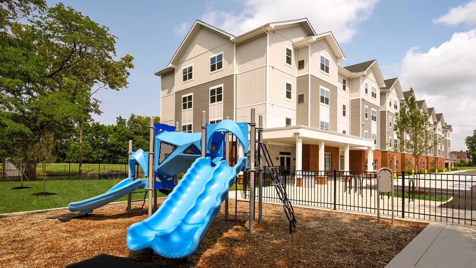 A playground with blue slides on wood mulch inside a black fence, with a four-story apartment building of beige siding and brick behind it