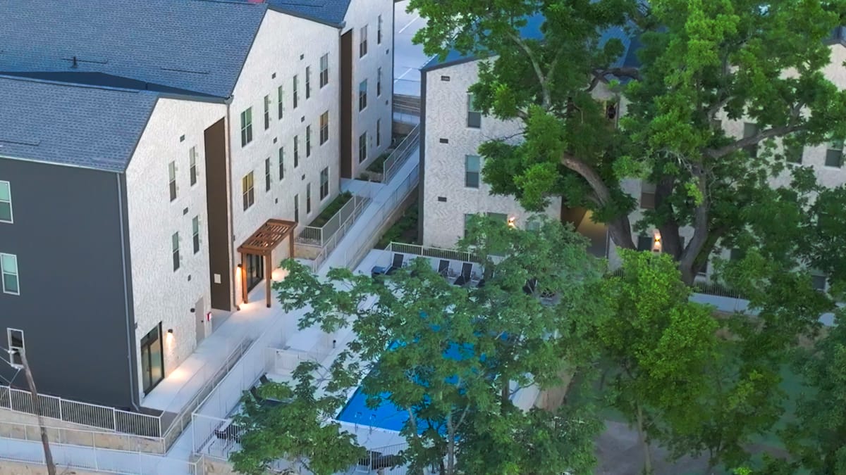 Aerial view at dusk of pale brick apartment buildings with dark roofs beside a swimming pool, partly hidden by large trees