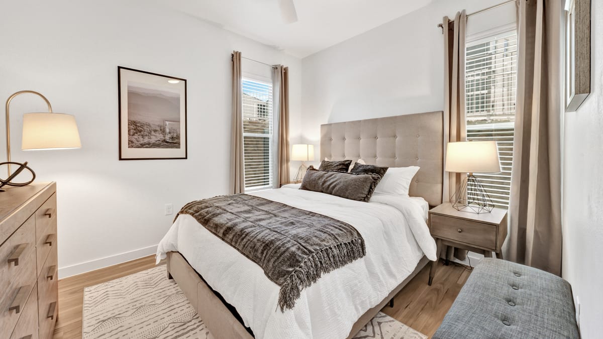 Bedroom with an upholstered bed, two windows with curtains, a ceiling fan, a dresser and wood-style flooring