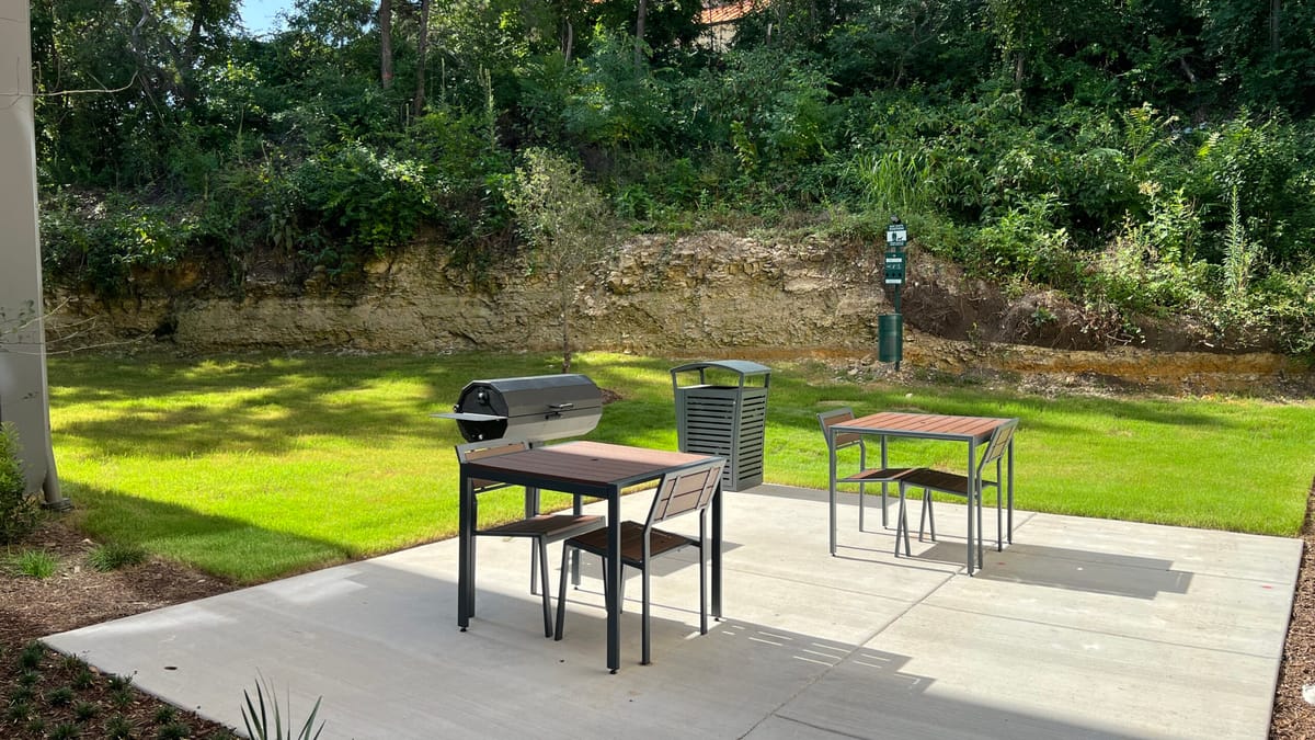 Concrete patio with a barrel grill, two metal tables with chairs and a lawn below a wooded limestone bluff