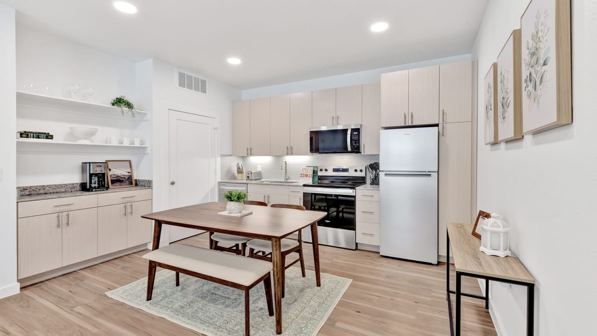 Open kitchen with pale flat-front cabinets, a white refrigerator, a stainless steel range and microwave, and a wooden dining table with a bench