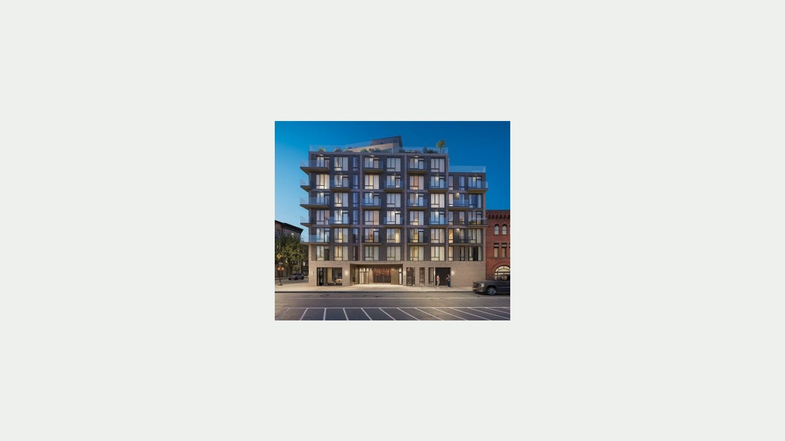 Rendering at dusk of an eight-story apartment building faced in dark gray brick with light vertical bands, glass balconies at the corners and a set-back roof level with plants, a pale stone ground floor with a lit lobby entrance, a red-brick building at right, a parked pickup truck and a striped parking lane in the foreground