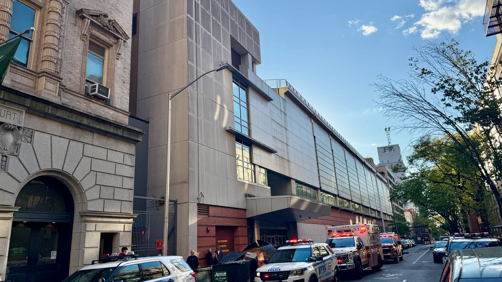 Photograph of West 54th Street with a tall windowless gray-paneled building and a long low gray building behind it, an ornate stone courthouse at left, a street lamp, dumpsters, and NYPD cars, an FDNY ambulance and other emergency vehicles parked along the curb under a blue sky