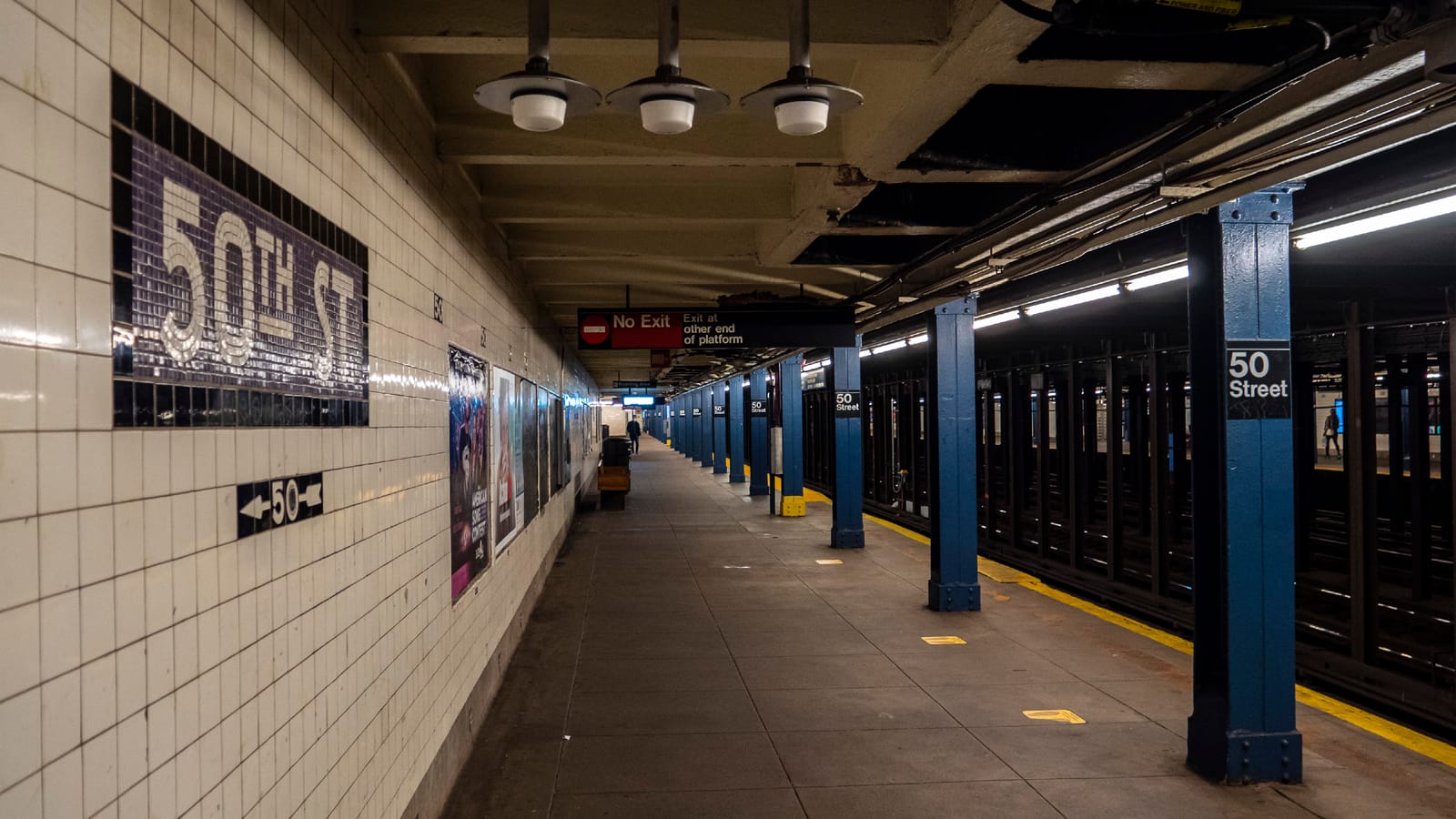 Photograph of an empty underground subway platform with white tiled walls, a black and white mosaic sign reading 50th St, blue steel columns marked 50 Street, ceiling lights and a red No Exit sign