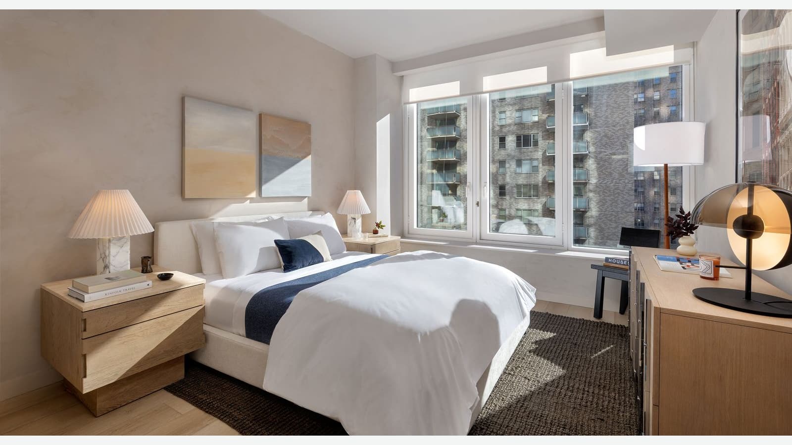 Photograph of a furnished bedroom with a white bed and blue throw, wood nightstands with pleated lamps, two paintings, a wide three-part window looking onto a brick apartment building with balconies, and a wood dresser with a lamp