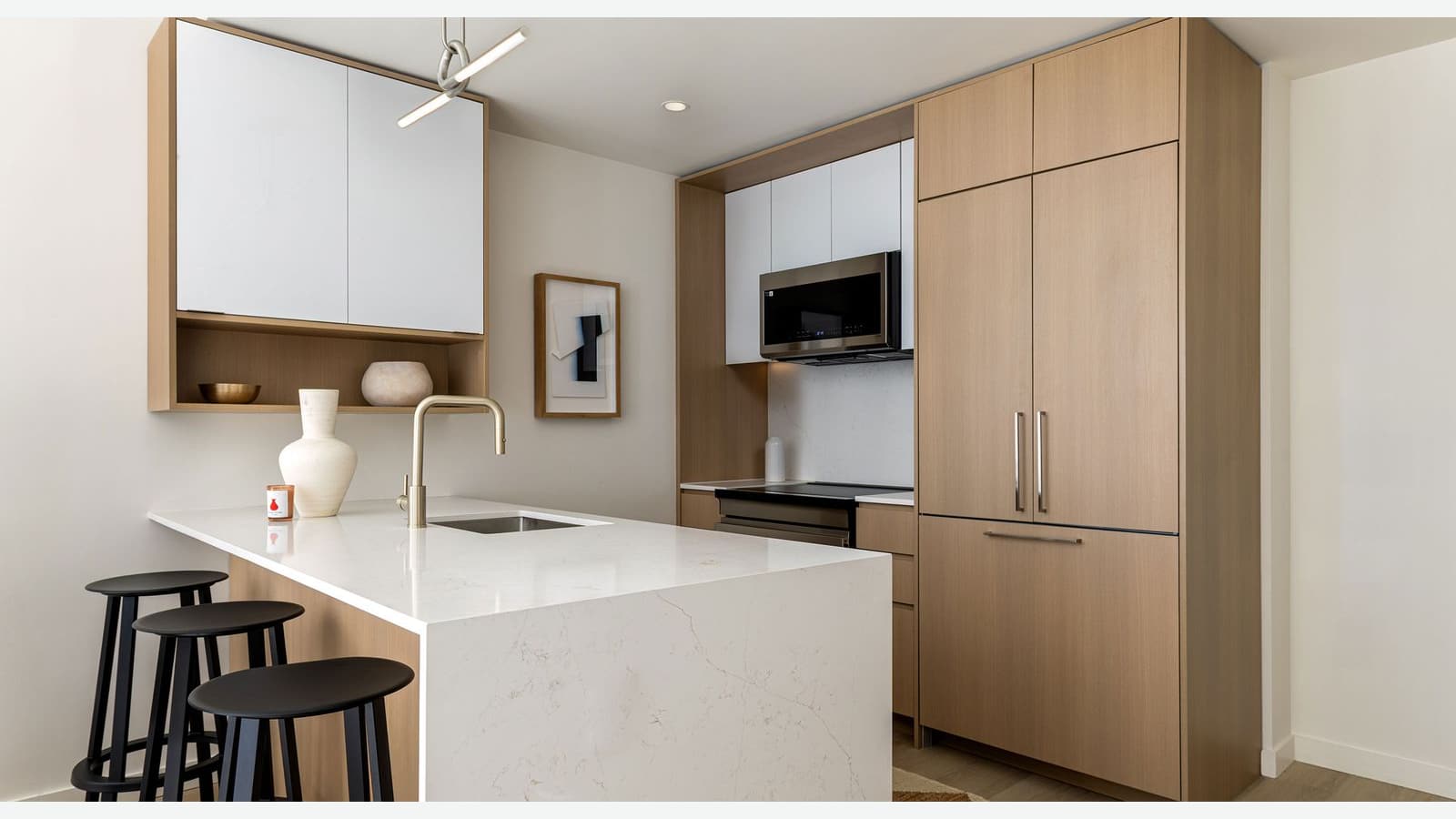 Photograph of a kitchen with a white stone peninsula and three black stools, a gold faucet, white and light wood cabinets, a microwave above a black glass electric cooktop and oven, and a tall wood-paneled refrigerator door