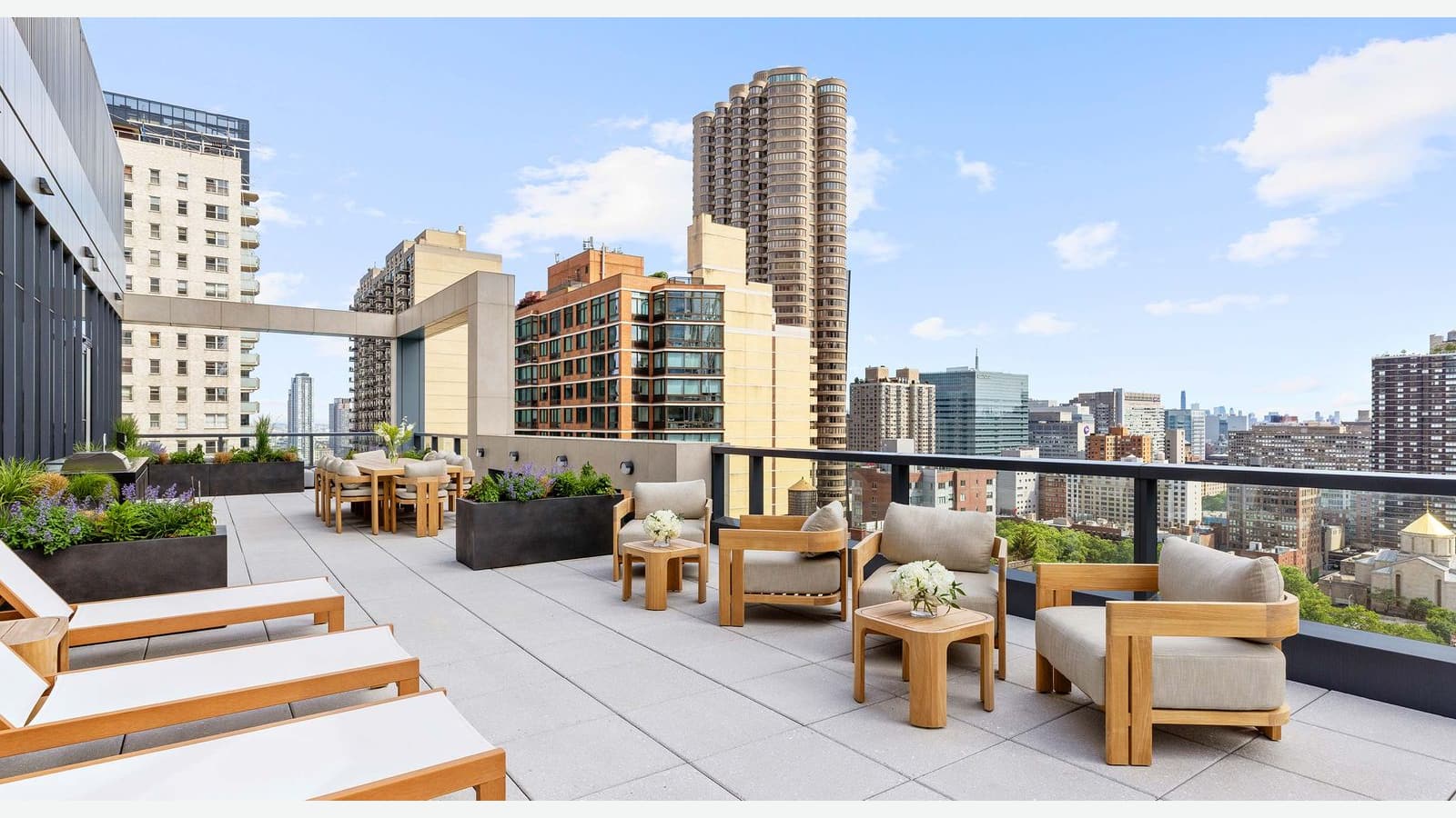 Photograph of a rooftop terrace with wood lounge chairs and loungers, planters of grasses and flowers, dining tables, a grill and a glass railing, with mid-rise and high-rise apartment towers and a round-edged tower beyond under a blue sky
