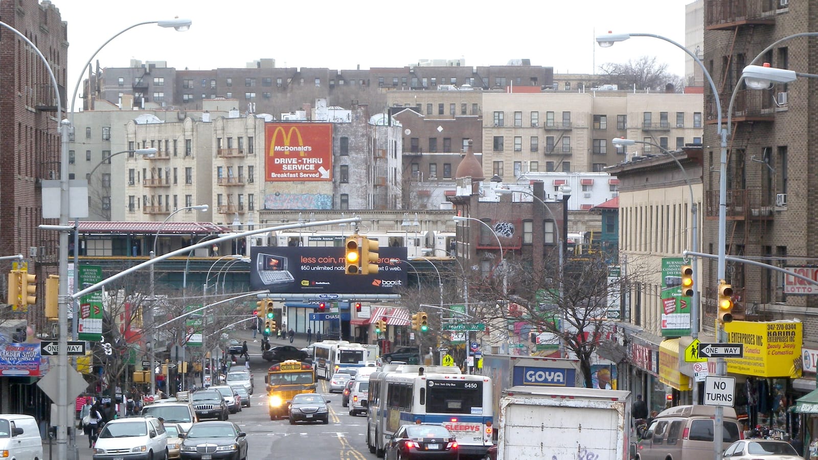 Photograph looking down a busy street lined with brick and stone apartment buildings and shops toward a raised subway station with a train standing in it, with billboards, traffic lights, a city bus, a yellow school bus and cars on a cloudy day