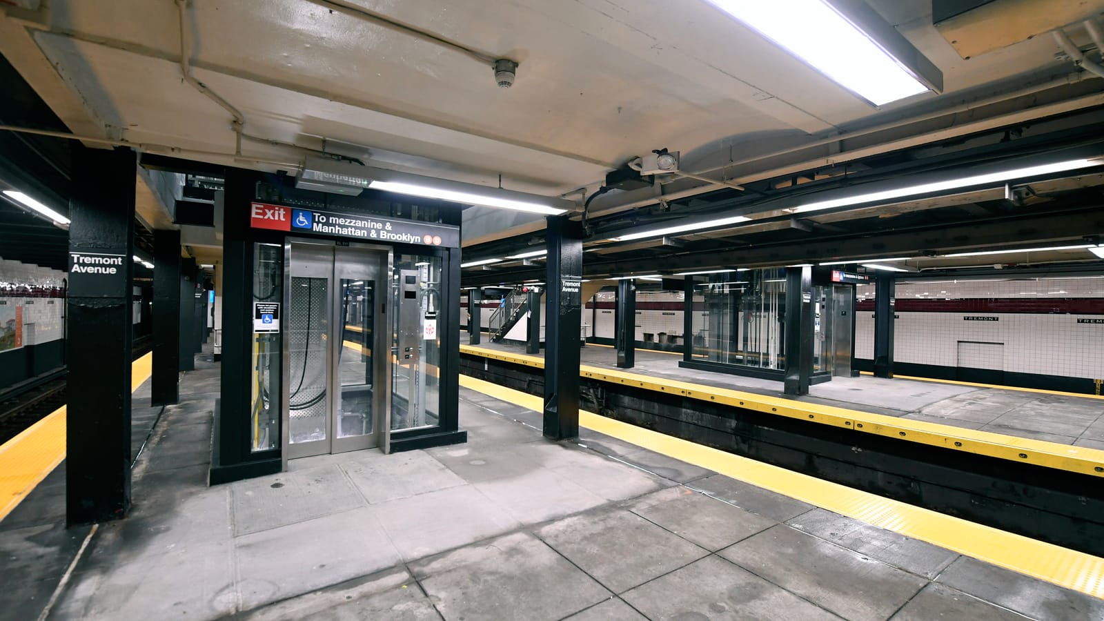 Photograph of an empty subway platform with a glass and steel elevator under a sign reading Exit, a wheelchair symbol and To mezzanine and Manhattan and Brooklyn, black columns marked Tremont Avenue, yellow platform edges and a second elevator across the tracks