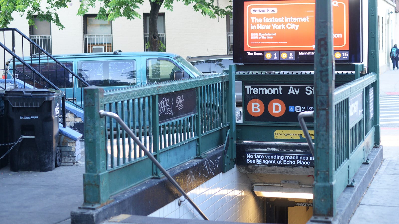 Photograph of a green iron subway stair entrance with globe lamps and a sign reading Tremont Av St with orange B and D circles and wheelchair and elevator symbols, an ad screen above it, a parked teal van and trash bins behind the railing