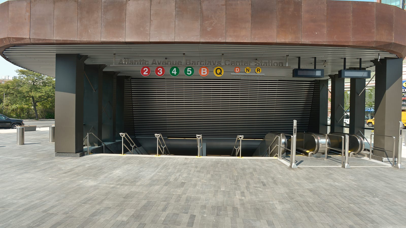 Photograph of a subway entrance under a wide curved canopy clad in rust-colored metal, with stairs and two escalators going down, the sign Atlantic Avenue Barclays Center Station with the 2, 3, 4, 5, B and Q symbols and D, N and R via passage, on a paved plaza with no people