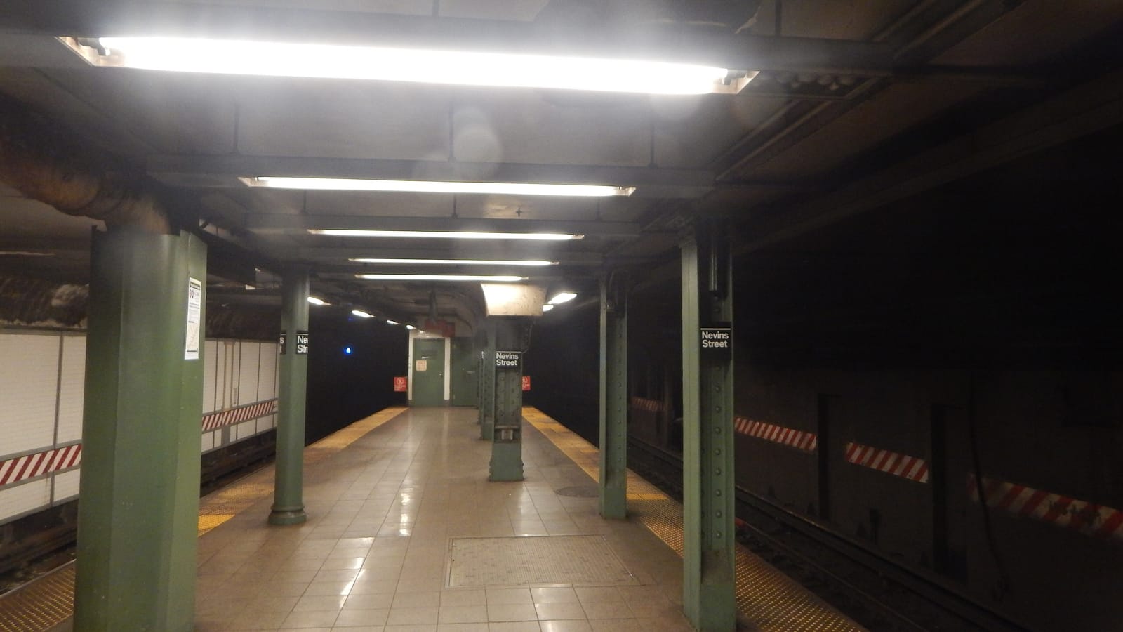 Photograph of an empty underground subway platform with a tiled floor, green steel columns with Nevins Street signs, fluorescent ceiling lights and tracks along both sides