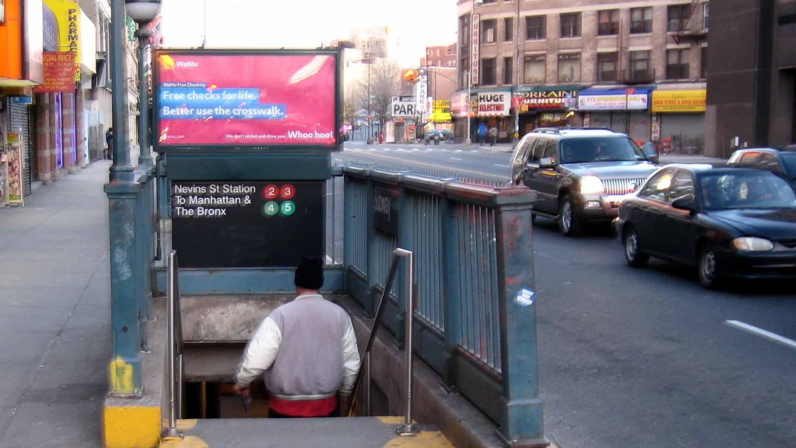 Photograph of a green iron subway stair entrance on a wide avenue sidewalk, with a sign reading Nevins St Station To Manhattan and The Bronx and the 2, 3, 4 and 5 symbols, a man in a jacket seen from behind walking down the steps, cars on the avenue and shop fronts across the street