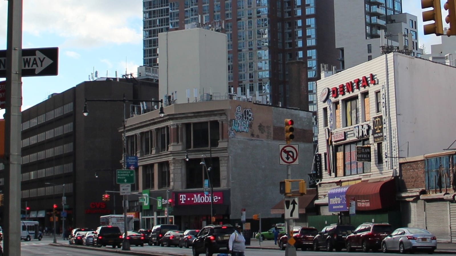 Photograph of a street corner on Flatbush Avenue with a four-story stone-trimmed building holding a T-Mobile store, a white building with Dental and Podiatrist signs, parked cars and a pedestrian, and behind them a tall glass and red-brick apartment tower against a cloudy sky