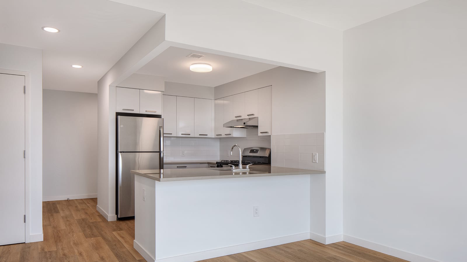Empty apartment with a white open kitchen: flat white cabinets, a stainless steel refrigerator and range, a grey counter with a sink on a half wall, and wood-look plank flooring