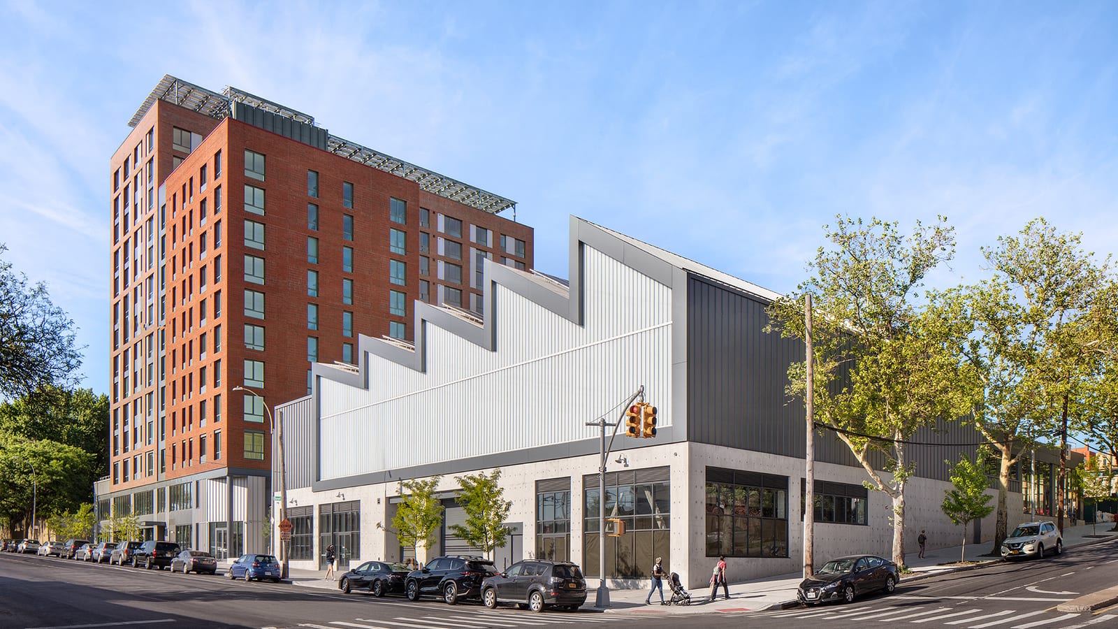 Photograph of a red brick apartment building of about fourteen stories with solar panels on the roof, behind a low grey and white building with a sawtooth roofline on a street corner, with parked cars and a person pushing a stroller