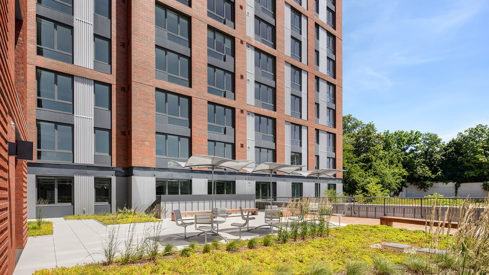 Photograph of a paved terrace with metal tables, chairs and shade canopies beside planted beds, at the base of a red brick apartment building with grey metal panels between the windows