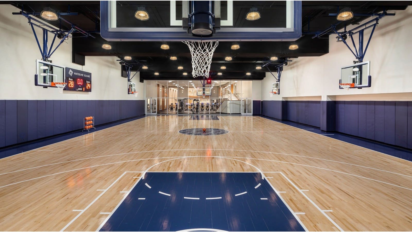 An indoor basketball court with a maple floor and navy paint, a hoop hanging overhead in the foreground, side hoops on the walls, a scoreboard reading Waterline Square and a glass wall opening onto a gym with treadmills