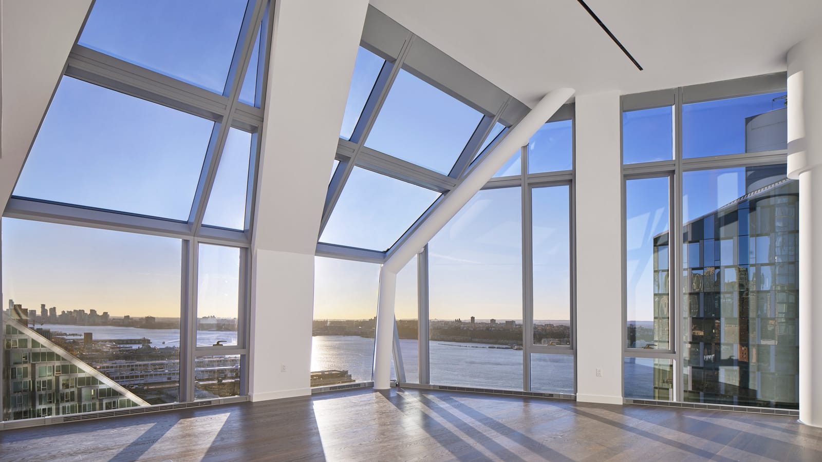 An empty, sunlit room with a dark wood floor and floor-to-ceiling windows, some of them slanting inward overhead, looking west over the Hudson River, with a white diagonal column and a glass tower at right