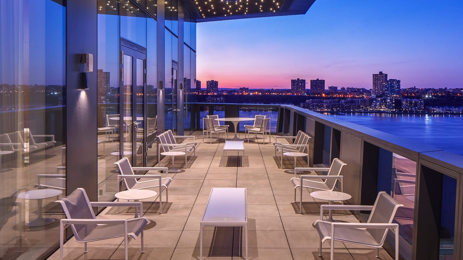An outdoor terrace at dusk with rows of white metal lounge chairs and low tables beside a glass wall, under a canopy studded with small lights, looking across the Hudson River to lit apartment towers on the far shore