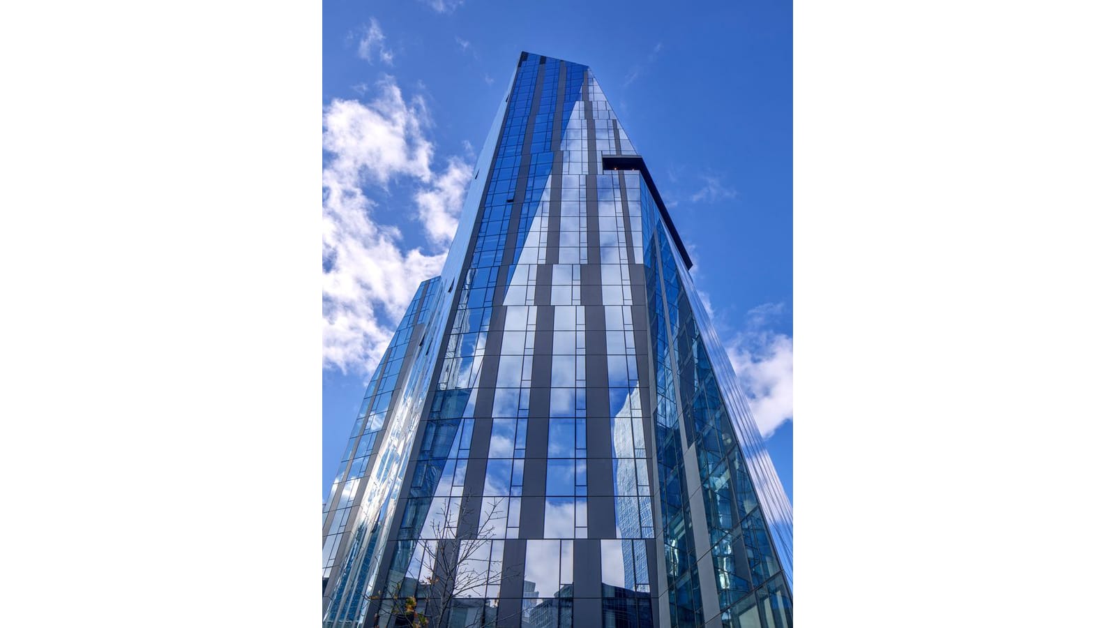 A faceted glass tower seen from street level looking straight up, its dark vertical mullions and angled glass planes reflecting blue sky and white clouds, with a bare tree at the bottom